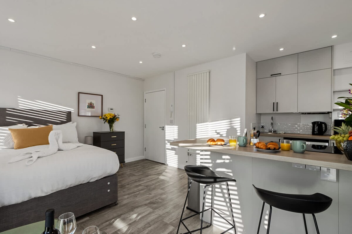 A modern studio apartment features a spacious bed adorned with neutral bedding and decorative pillows. The kitchenette is equipped with sleek cabinetry and appliances. Natural light filters through windows, casting striped shadows on the floor. A dining area with stools is set with cheerful tableware and fresh produce.