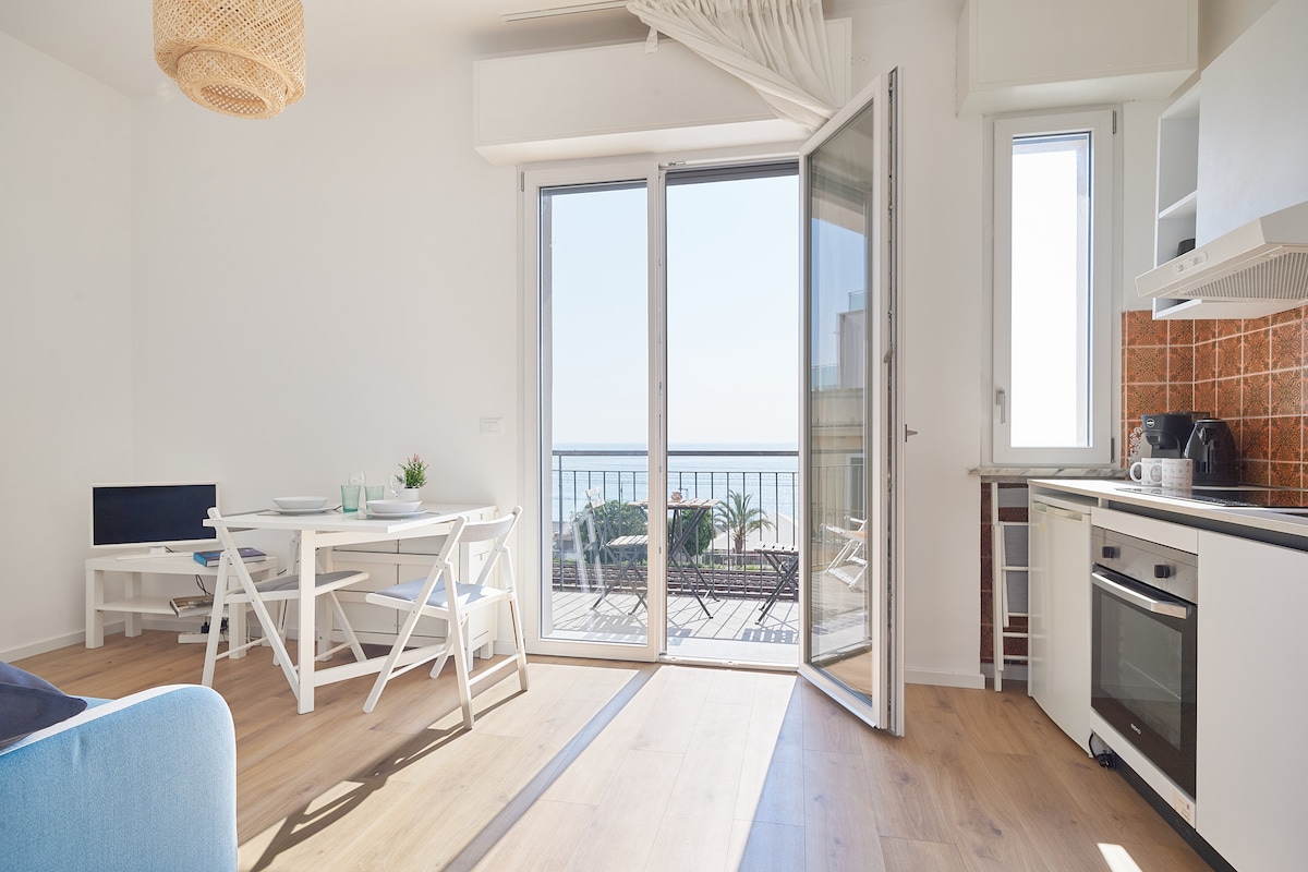 An open-plan living area features large glass doors that provide views of the sea. A white dining table and chairs are positioned beside the kitchen. A blue sofa offers comfortable seating. Natural light fills the space, enhancing the bright and airy atmosphere.