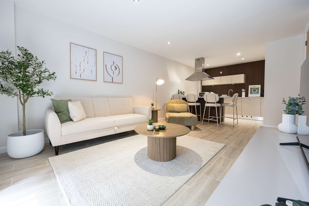A light-filled living area features a modern beige sofa positioned beside a decorative plant in a white pot. A round wooden coffee table is centered on a textured rug, complementing the contemporary kitchen visible in the background.