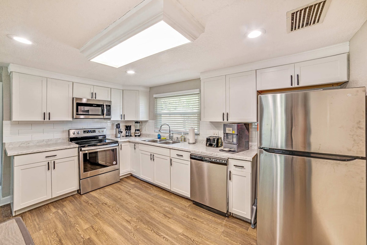 A modern kitchen is showcased with white cabinetry and stainless steel appliances, including a refrigerator and dishwasher. A spacious countertop provides ample workspace, while a window offers natural light. Essential kitchen items are organized neatly for convenience, enhancing the functional layout.