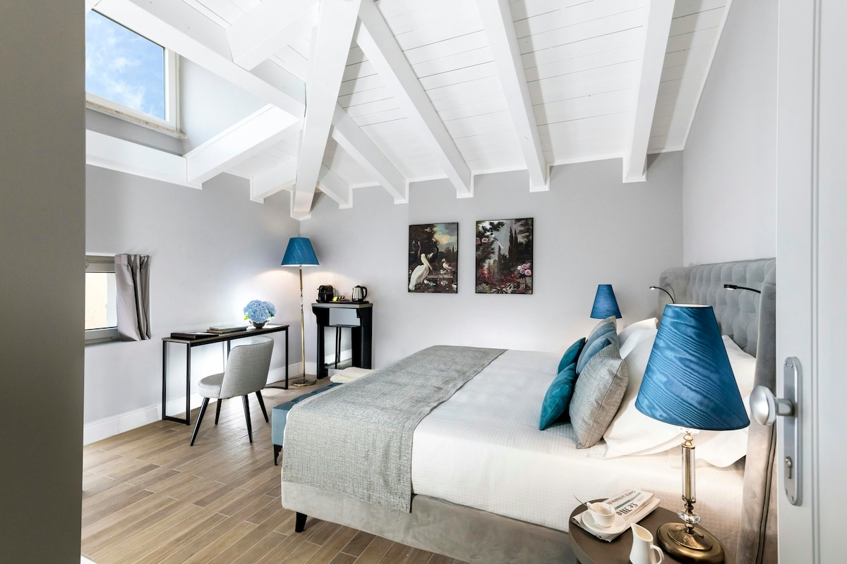 A well-lit room features a cozy double bed with soft linens and decorative pillows. Two blue lamps provide ambient lighting beside the bed. A desk with a chair is visible, along with a minibar and coffee maker. Art pieces adorn the walls, enhancing the modern design.