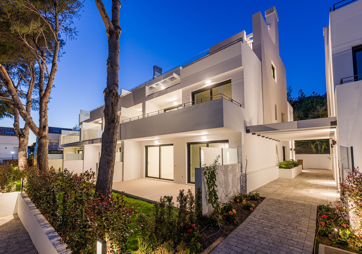 The modern building is situated in a well-maintained garden, showcasing wide balconies and large glass windows that reflect the evening sky. Pathways lead through lush greenery, enhancing the serene atmosphere of the surrounding area.