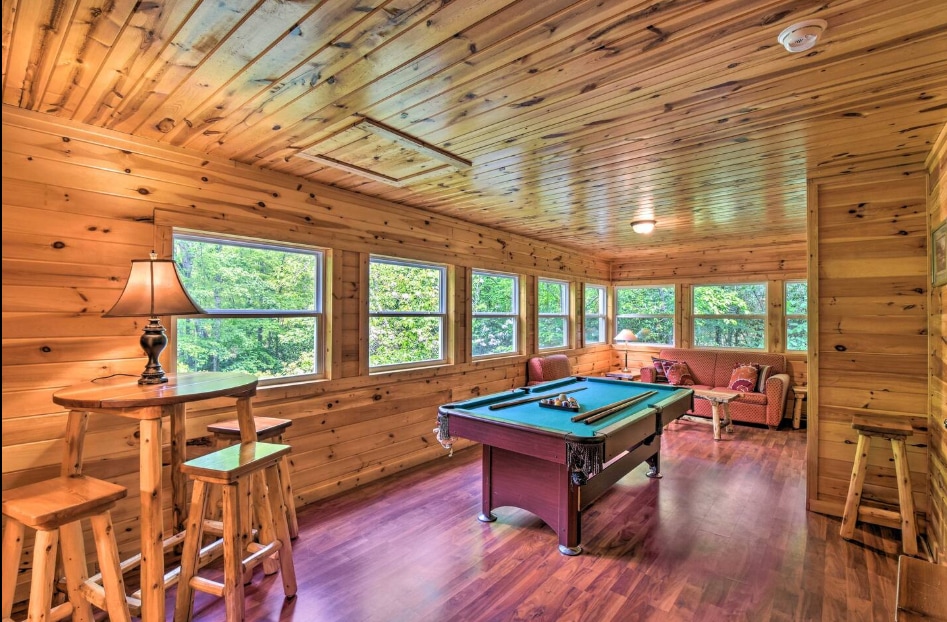 A spacious game room features a pool table and wooden accents throughout. Large windows allow natural light to fill the space, offering views of the surrounding greenery. A small dining table with stools and a comfortable seating area enhance the inviting atmosphere.
