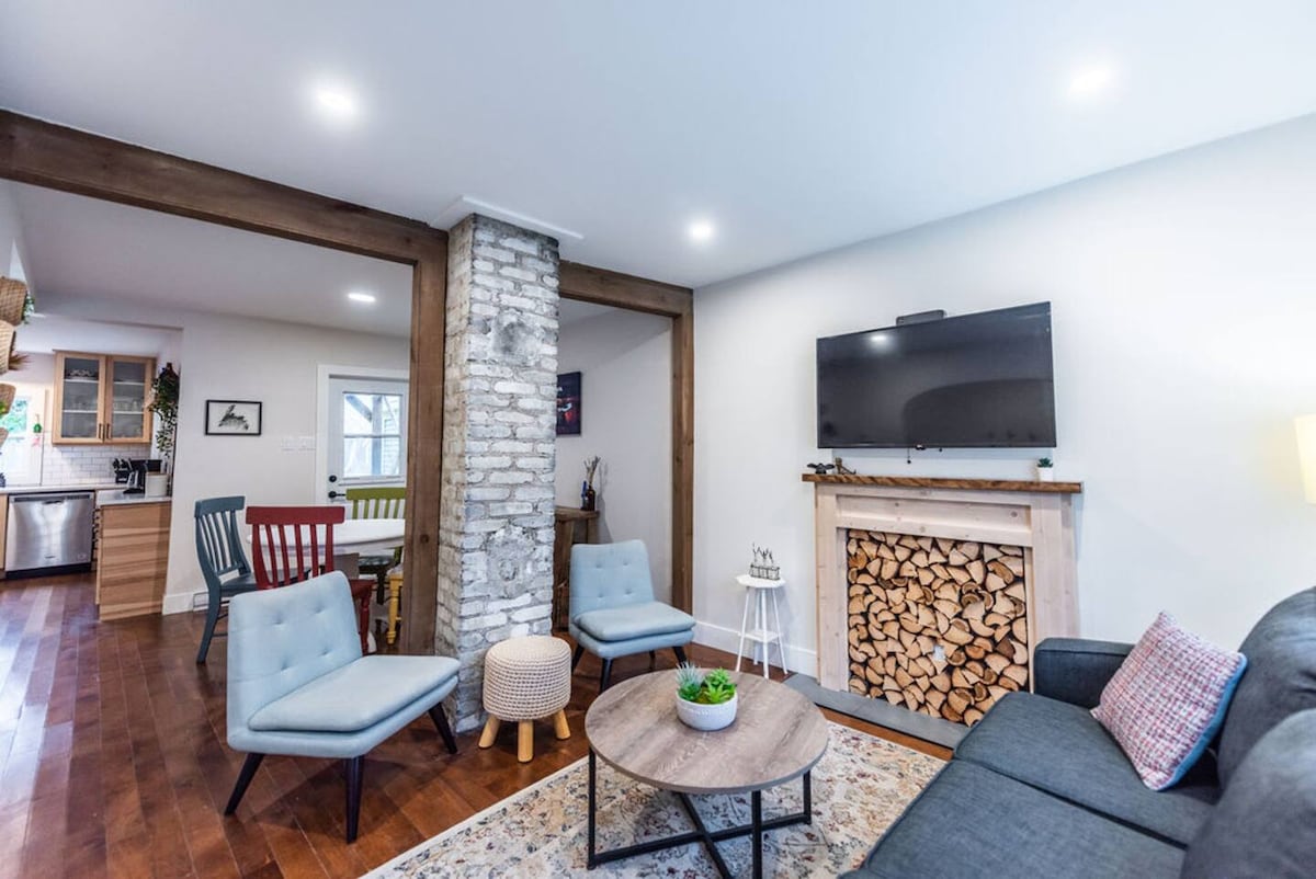 An open-concept living area is shown, featuring a modern design and warm wood accents. A cozy seating arrangement consists of three light-colored chairs and a dark sofa, with a circular table at the center. A stone fireplace, stacked with wood, adds character, and a mounted flat-screen TV completes the space.