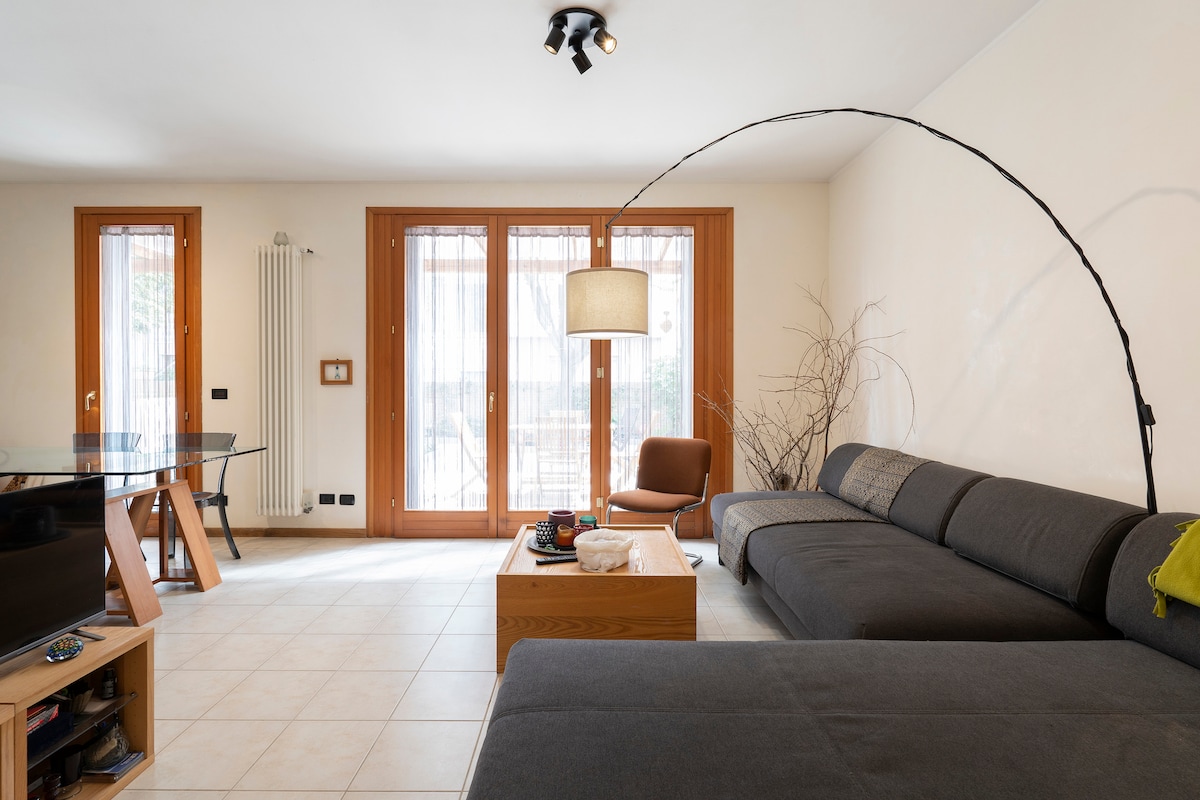 A spacious living room features large windows that invite natural light, highlighting the modern design. A cozy gray sectional sofa is paired with a wooden coffee table, while a glass dining table stands nearby. A tall floor lamp and decorative branches add the finishing touches.