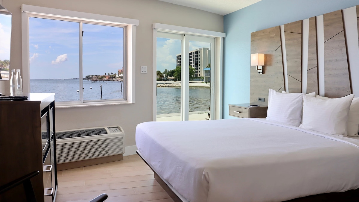 A spacious room features a large bed with crisp white linens. Natural light floods the area from a window and sliding glass door that offer views of the water and nearby marina. Minimalist furnishings include a nightstand and dresser.