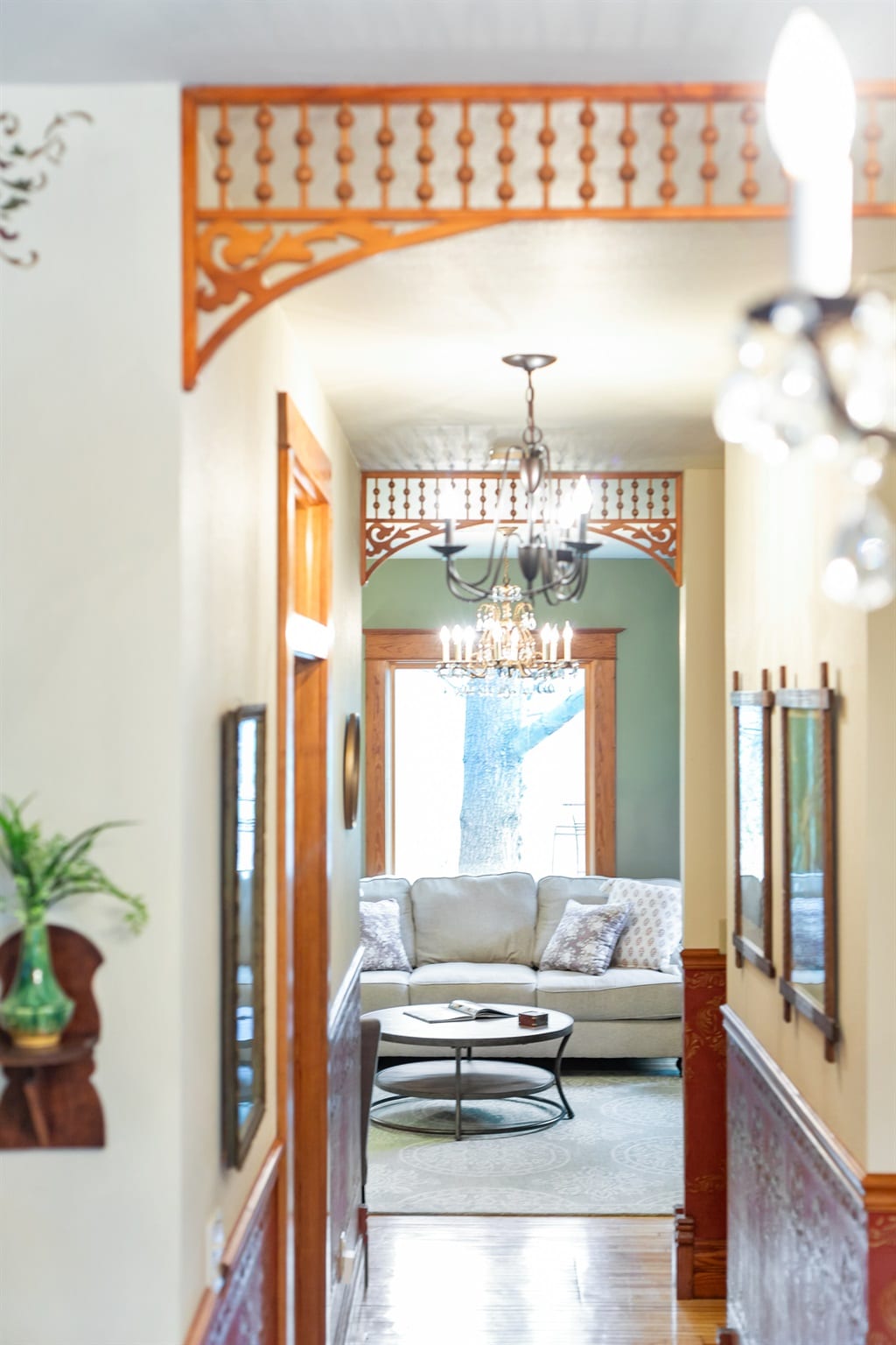 A hallway showcases detailed woodwork and ornate light fixtures, guiding the eye towards a cozy living area. Sunlight filters through large windows, illuminating a comfortable sofa layered with decorative pillows, positioned around a circular coffee table on a patterned rug.