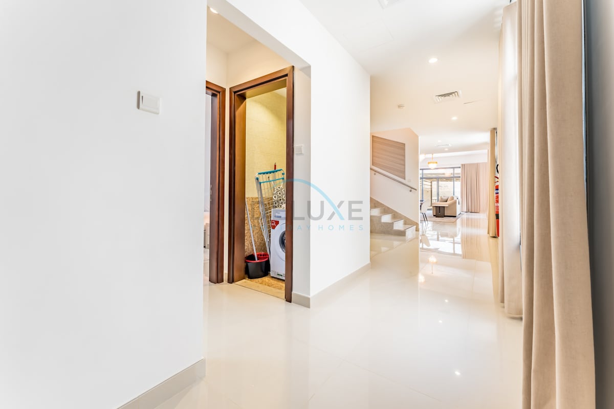 A tidy hallway is visible, illuminated by natural light. Doors on the left lead to a laundry room, while the wider space on the right transitions into a bright living area. The smooth, glossy floor reflects the light and enhances the open ambiance.