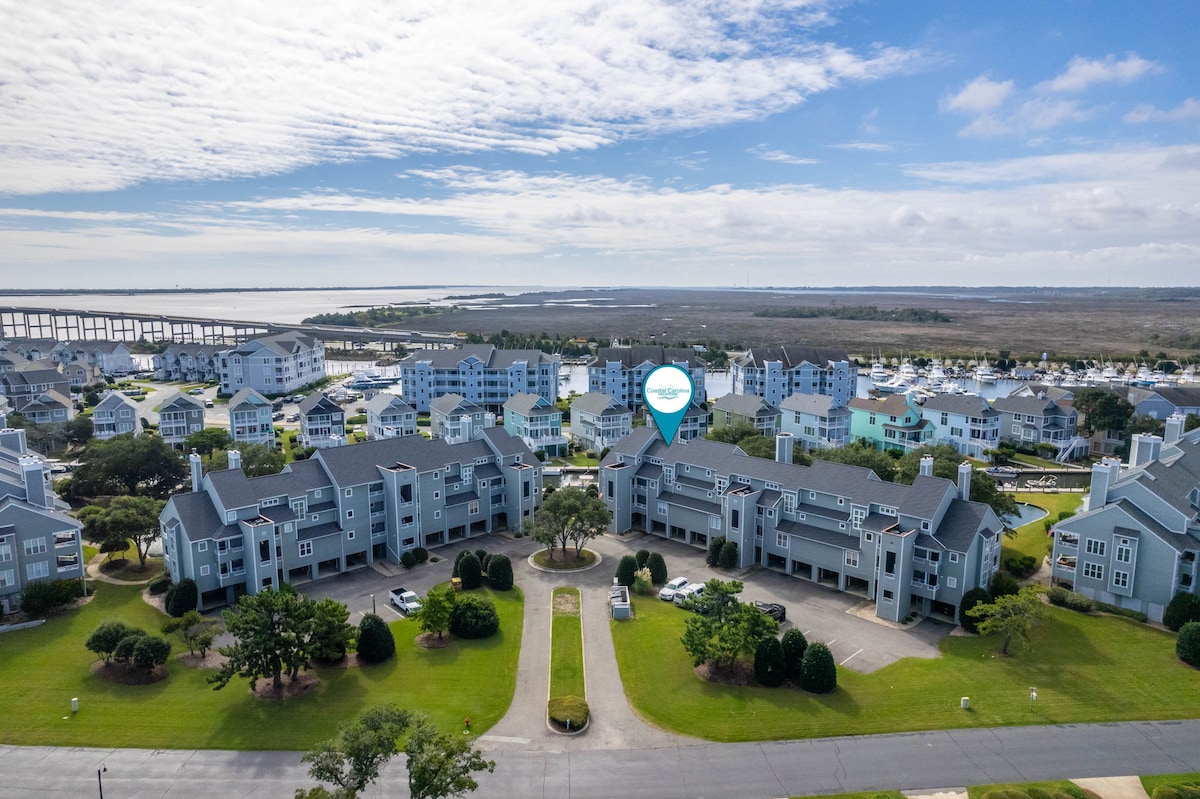 An aerial view captures the layout of the Pirate's Cove community, showcasing multiple two-story buildings surrounded by well-maintained lawns and trees. The nearby water is visible, reflecting the sky, while the Washington Baum bridge spans in the distance.