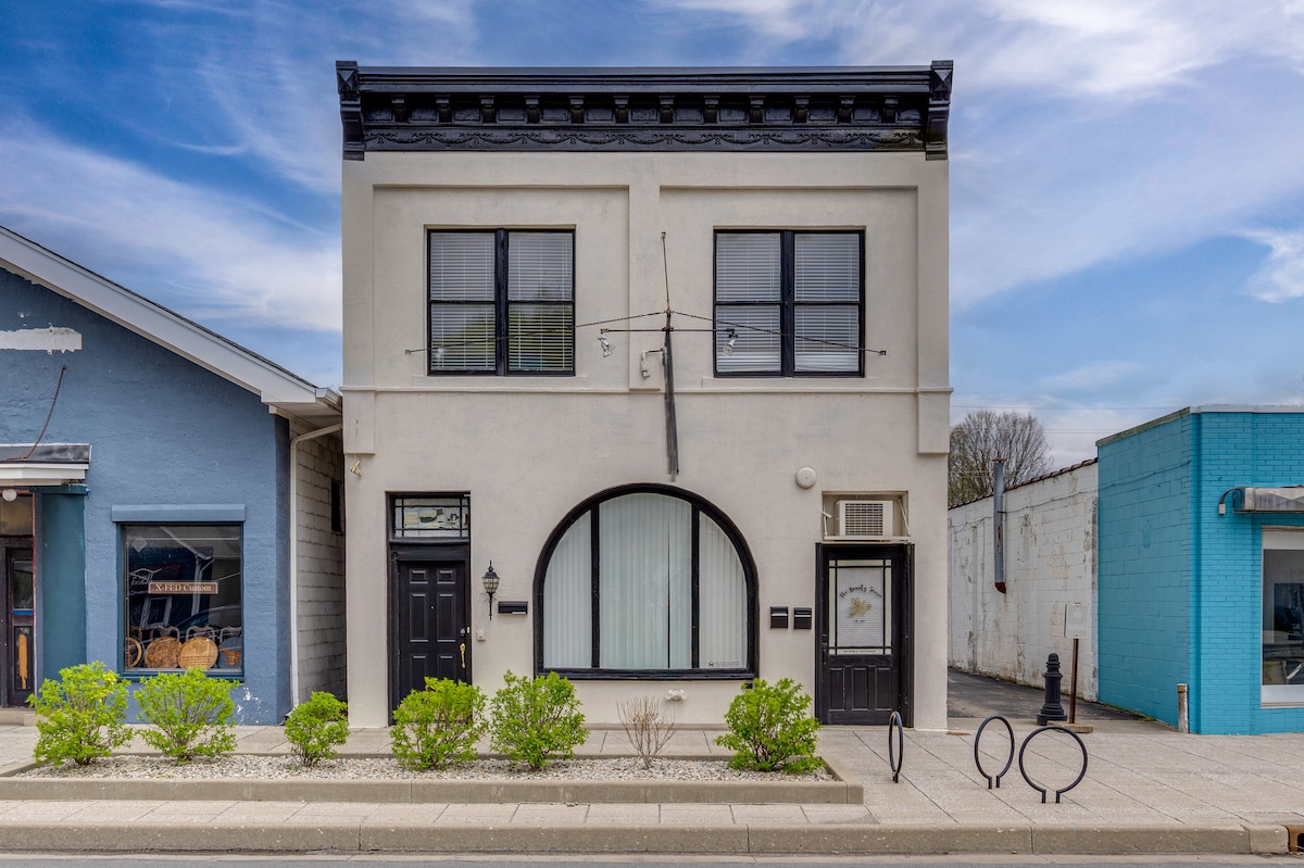 The exterior of a two-story building is presented, featuring a smooth white façade with black accents. Large windows are framed stylishly, with an arched window detail creating a focal point. Lush green shrubs line the walkway, complemented by clear blue skies overhead.