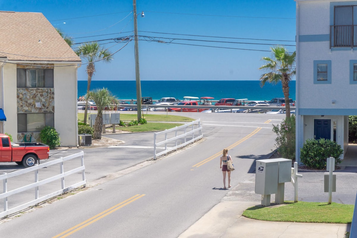 A clear view highlights the street leading to the beach, with a sunny blue sky and the ocean visible in the distance. Palm trees border the road, and parked vehicles line the opposite side, suggesting easy beach access just across the street.