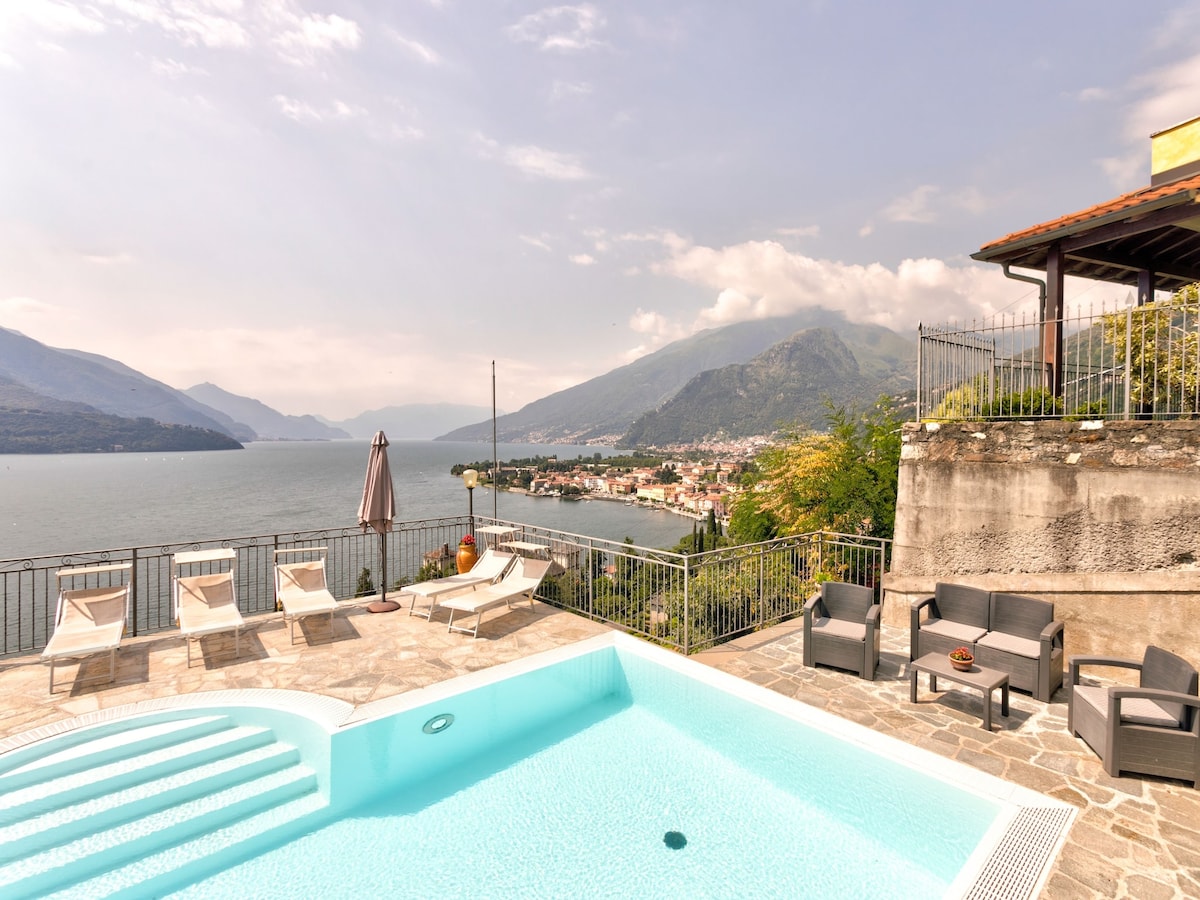 A shared swimming pool is visible, featuring light blue water and steps leading in. Surrounding the pool are several sun loungers and seating areas, with scenic views of the lake and mountains in the background. The area is framed by a stone wall and greenery.