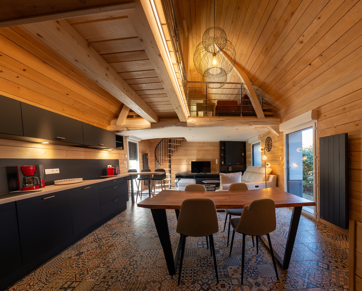 The kitchen and dining area feature a wooden structure with high ceilings, illuminated by pendant lighting. A large table is surrounded by four chairs, while modern kitchen cabinets offer ample storage. A staircase leads to the upper level, providing additional living space.