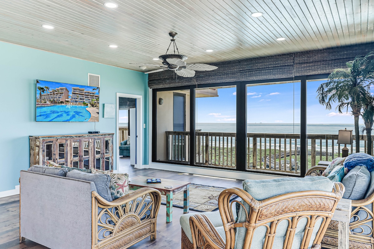 A spacious living area is highlighted by large floor-to-ceiling windows, offering panoramic views of the Gulf of Mexico. Comfortable seating arrangements, including a stylish couch and accent chairs, complement the decor. A television is mounted on the wall, enhancing relaxation opportunities.