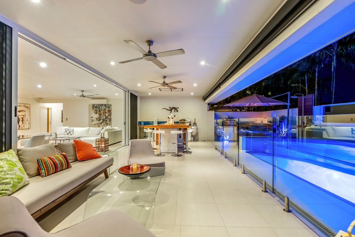 An open-plan living area is showcased, featuring contemporary furnishings and large glass sliding doors. The stylish space provides a view of the swim spa pool, complemented by ambient lighting. Ceiling fans are positioned overhead, and sleek tile flooring enhances the modern design.