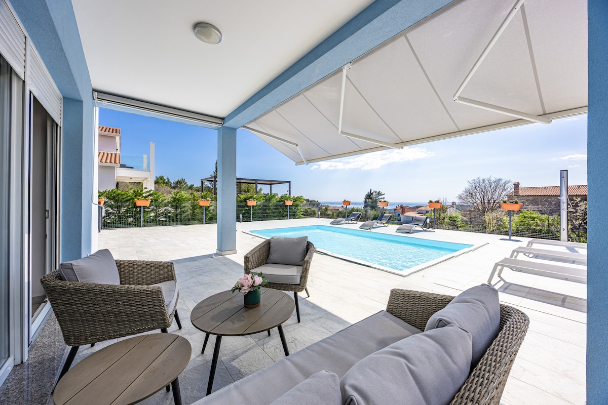 A covered outdoor seating area features comfortable wicker chairs surrounding a small table. The space overlooks a spacious pool area, showcasing sunbeds and a beautifully landscaped garden, with panoramic views of the surrounding landscape and clear skies.