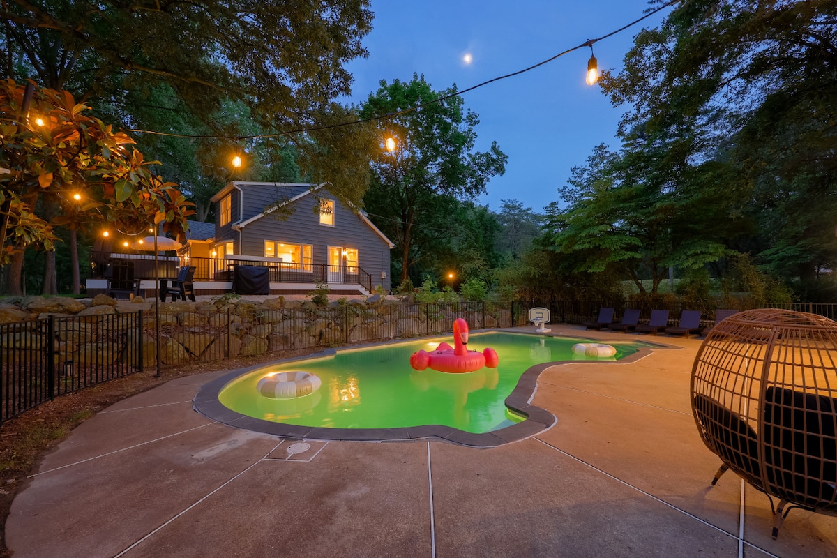 The heated saltwater pool is surrounded by a decorative fence, with charming outdoor lighting strung overhead. A pink inflatable float is visible in the water, alongside chic loungers positioned around the pool area. The peaceful backdrop includes trees and the newly renovated cottage.