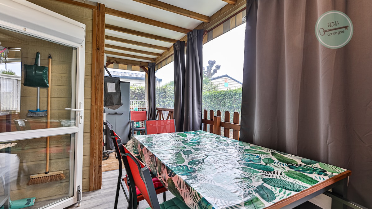 A covered terrace area is visible, featuring a spacious dining table with a leaf-patterned tablecloth. Several red chairs surround the table. Large windows are lined with curtains, providing a view of the surrounding greenery and residential space.