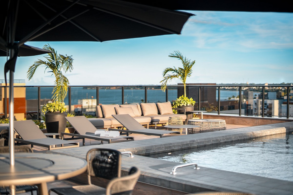 A rooftop pool area is presented with sun loungers arranged around the water's edge. Comfortable seating is positioned nearby, offering views of the ocean and surrounding landscape. Potted plants and palm trees add greenery to the space, enhancing its outdoor appeal.