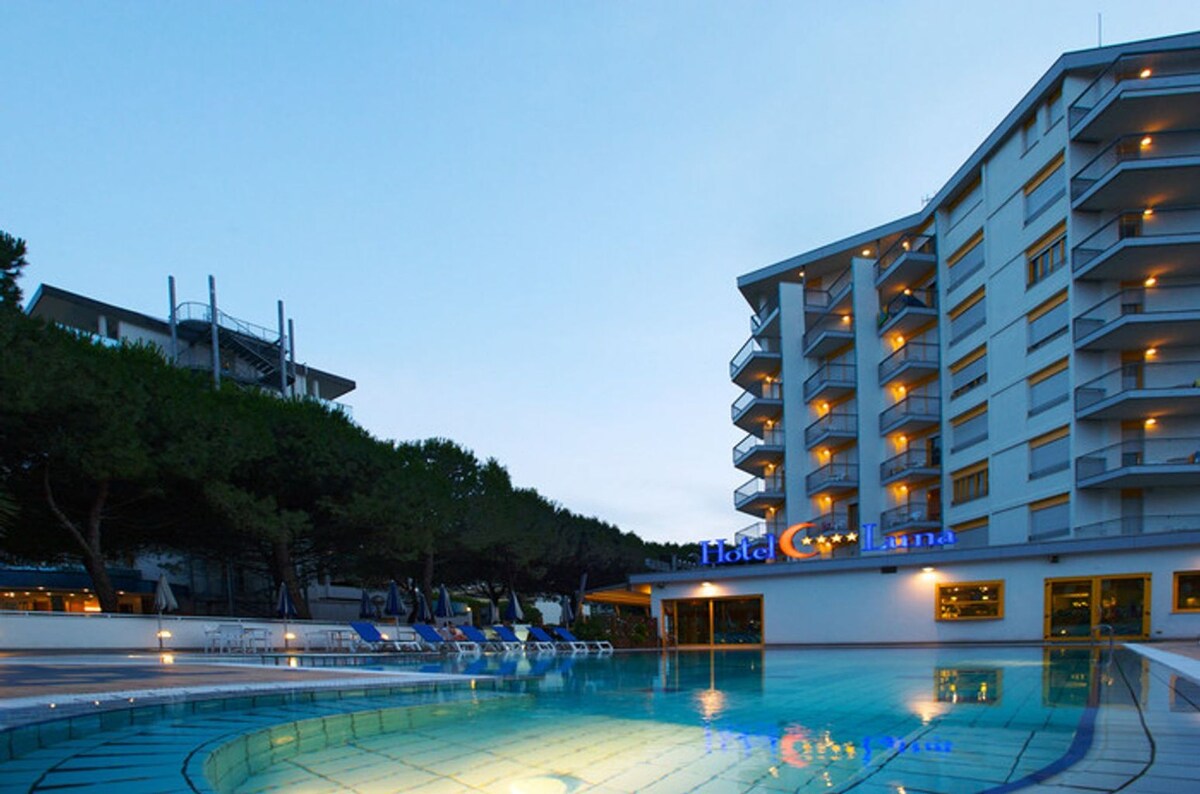 The exterior of Hotel Luna is displayed, featuring a multi-story building adjacent to a tranquil swimming pool. Comfortable loungers are positioned around the pool, surrounded by lush greenery. The hotel name is illuminated, creating a welcoming ambiance during the twilight hours.