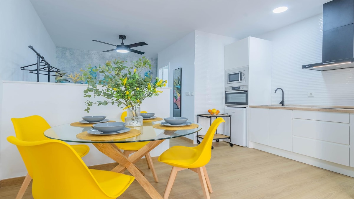 A bright dining area is presented with a glass table surrounded by four yellow chairs. A kitchen area features white cabinetry, a microwave, and a ceramic hob. A fan hangs from the ceiling, while decorative touches, including a vase with greenery, add to the inviting atmosphere.