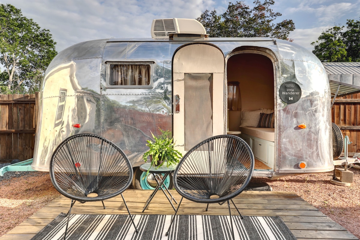 The exterior of a restored vintage Airstream is shown, featuring a shiny aluminum surface. A small entrance is visible alongside two black chairs on a wooden deck, adorned with a striped rug. Lush greenery surrounds the space, promoting a relaxing atmosphere.