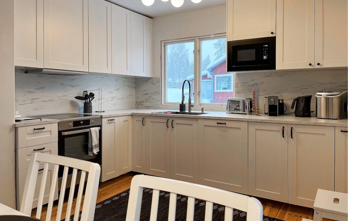 A modern kitchen is complemented by white cabinetry and light marble countertops. Natural light enters through a window overlooking a snowy outdoor scene. Appliances include an oven, microwave, and a coffee maker, while a dining table with white chairs sits invitingly nearby.