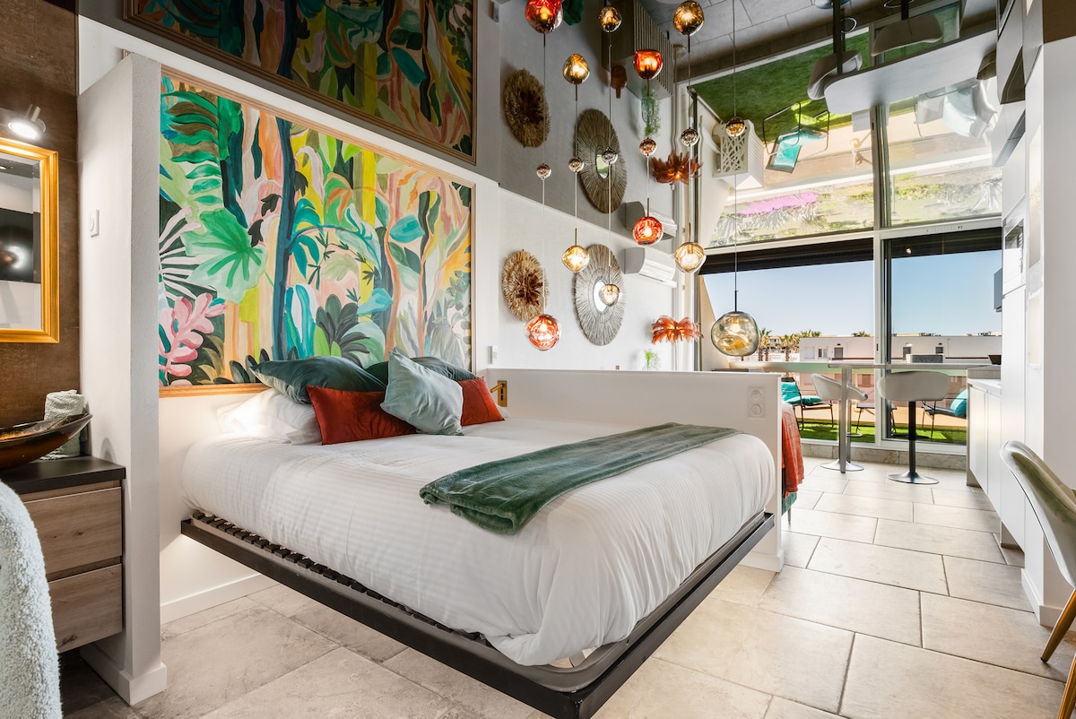 A vibrant sleeping area features a spacious bed adorned with colorful pillows. An artistic mural of lush greenery adds visual interest. Unique hanging light fixtures create a dynamic atmosphere, while large windows frame views of the outdoor terrace and the surroundings.