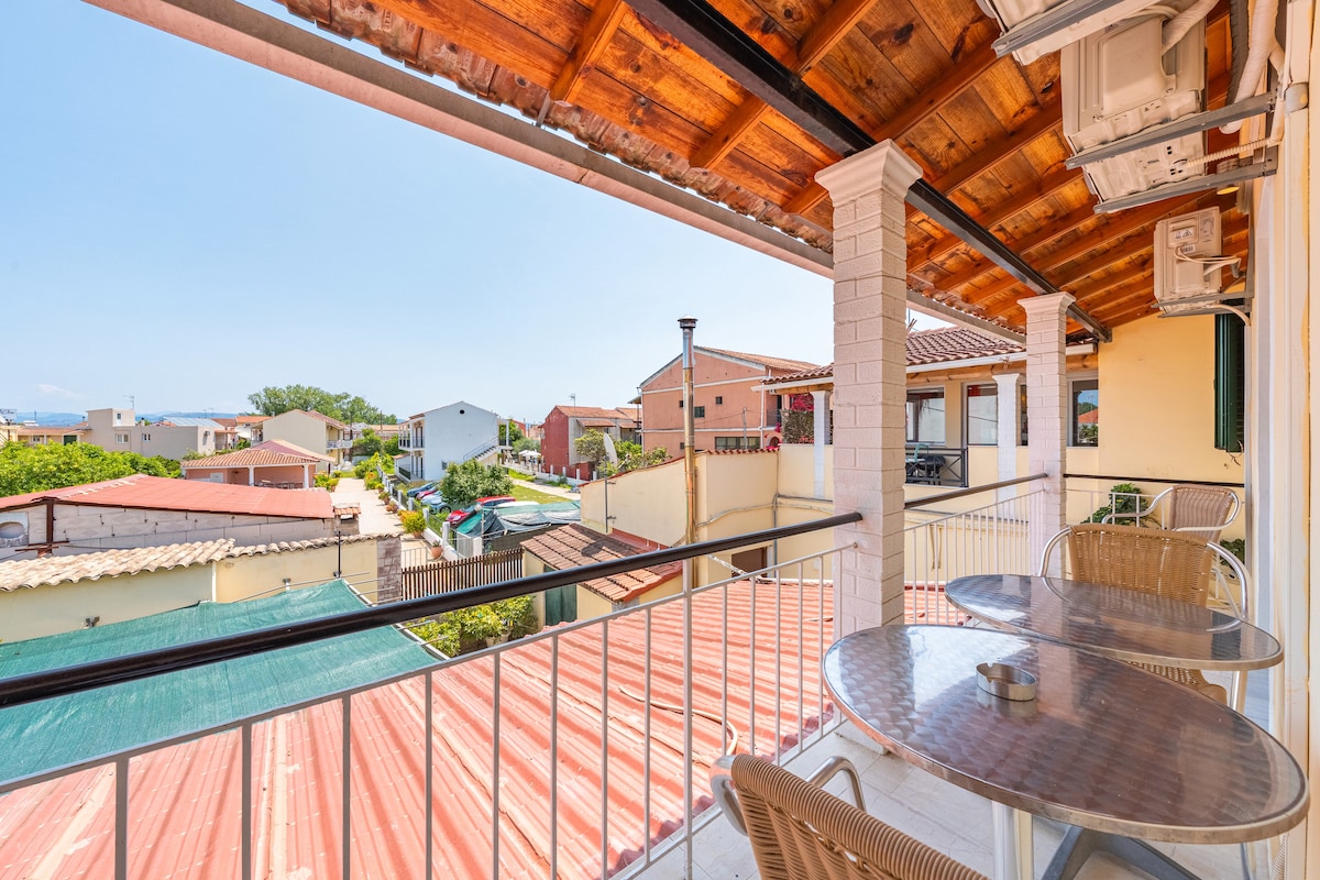 A covered balcony features a wooden ceiling and overlooks nearby rooftops. Two round tables with chairs are positioned on the balcony, providing a space for outdoor relaxation. The scenic view includes neighboring structures and partial glimpses of the surrounding area.