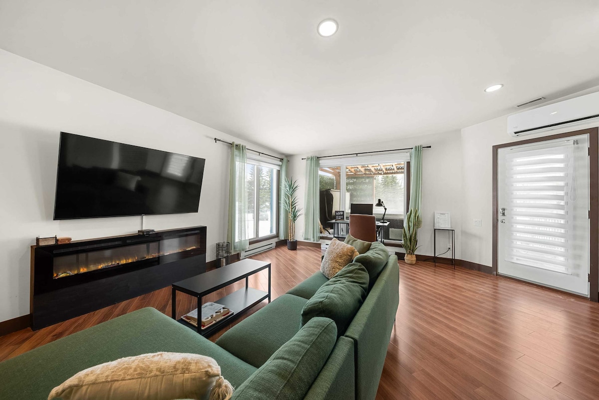 A spacious lounge area features a green sectional sofa, complemented by a coffee table. A large, wall-mounted television is positioned above a linear electric fireplace. Natural light enters through large windows, partially covered by sheer curtains, enhancing the welcoming atmosphere.