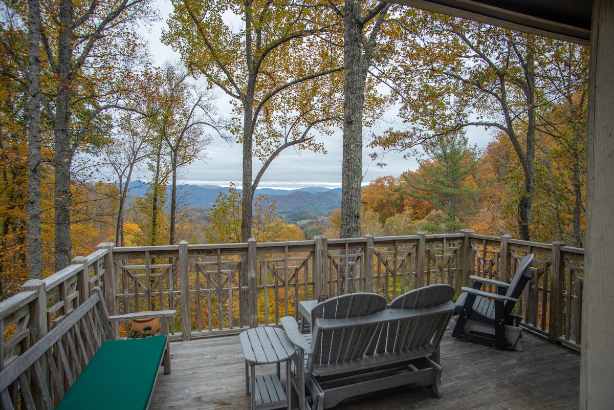 A spacious deck is surrounded by trees displaying fall foliage, providing a serene vista of the mountains in the distance. Comfortable wooden seating arrangements invite relaxation while taking in the scenic views.