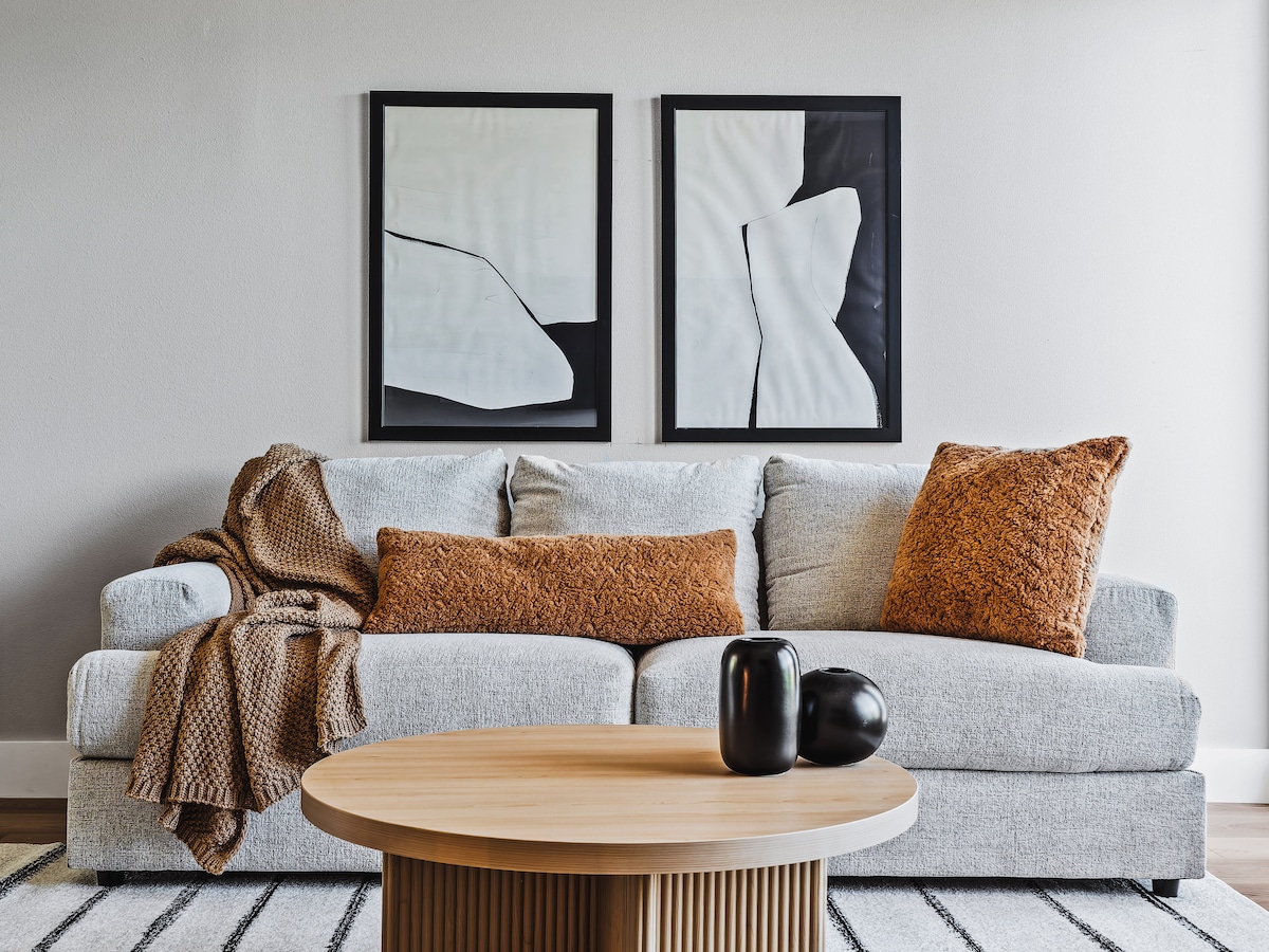 A modern living space features a light gray sofa adorned with textured pillows and a soft throw. Two minimalist wall artworks in black and white provide a stylish backdrop. A round wooden coffee table sits centered in front of the sofa, enhancing the relaxed ambiance.