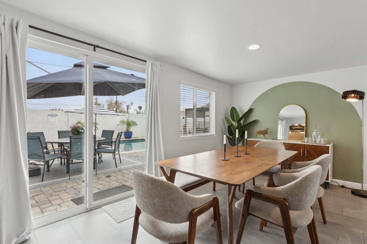 A dining area is presented with a warm wooden table surrounded by four upholstered chairs. Natural light filters through large sliding glass doors, leading to a patio with an umbrella and outdoor seating, while a green accent wall adds a modern touch.