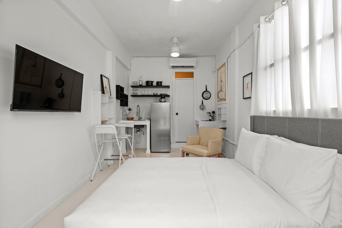 A cozy studio apartment features a spacious bed with white linens and soft pillows. A flat-screen TV is mounted on a light-colored wall. The kitchenette is equipped with essential appliances, and a small dining area with two white chairs is arranged nearby. Natural light filters through sheer curtains.