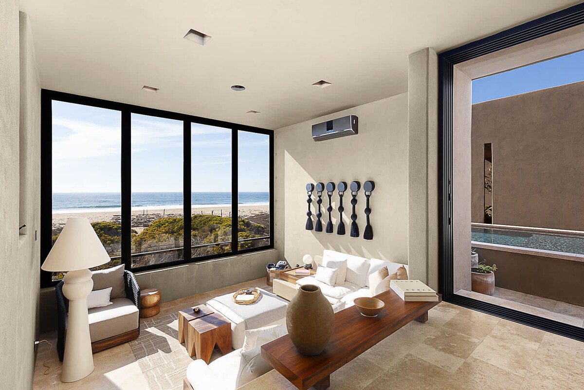 A spacious living area is highlighted by large windows that provide panoramic ocean views. The neutral-toned furniture includes a low wooden coffee table and white seating. Natural light illuminates the space, creating a bright and airy atmosphere, complemented by a decorative wall piece.
