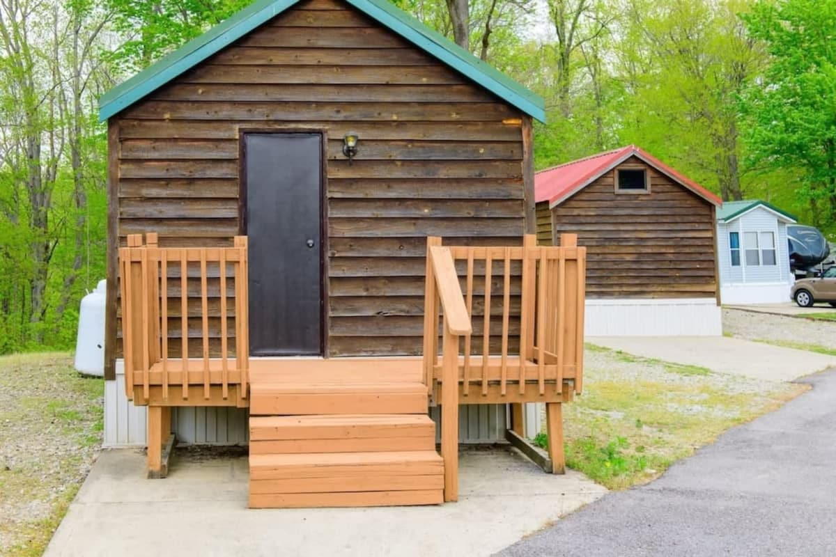 A cozy cabin exterior features wooden siding and a welcoming front door. A set of wooden steps leads to the entrance, flanked by a porch area, with additional cabins visible in the background. Lush greenery surrounds the cabin, highlighting the serene atmosphere.