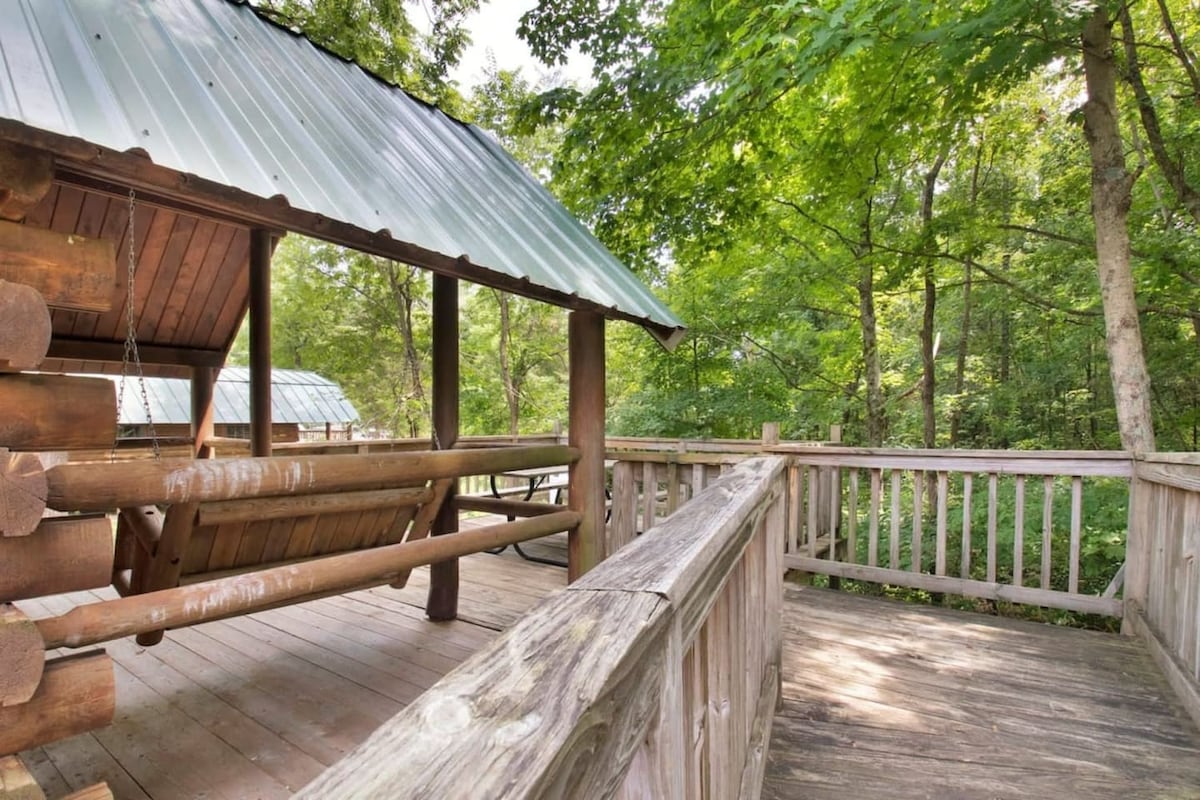 A wooden terrace is showcased with a rustic swing and seating area, surrounded by a lush green landscape. The structure features a sloped metal roof, and trees provide natural shade, enhancing the peaceful atmosphere.