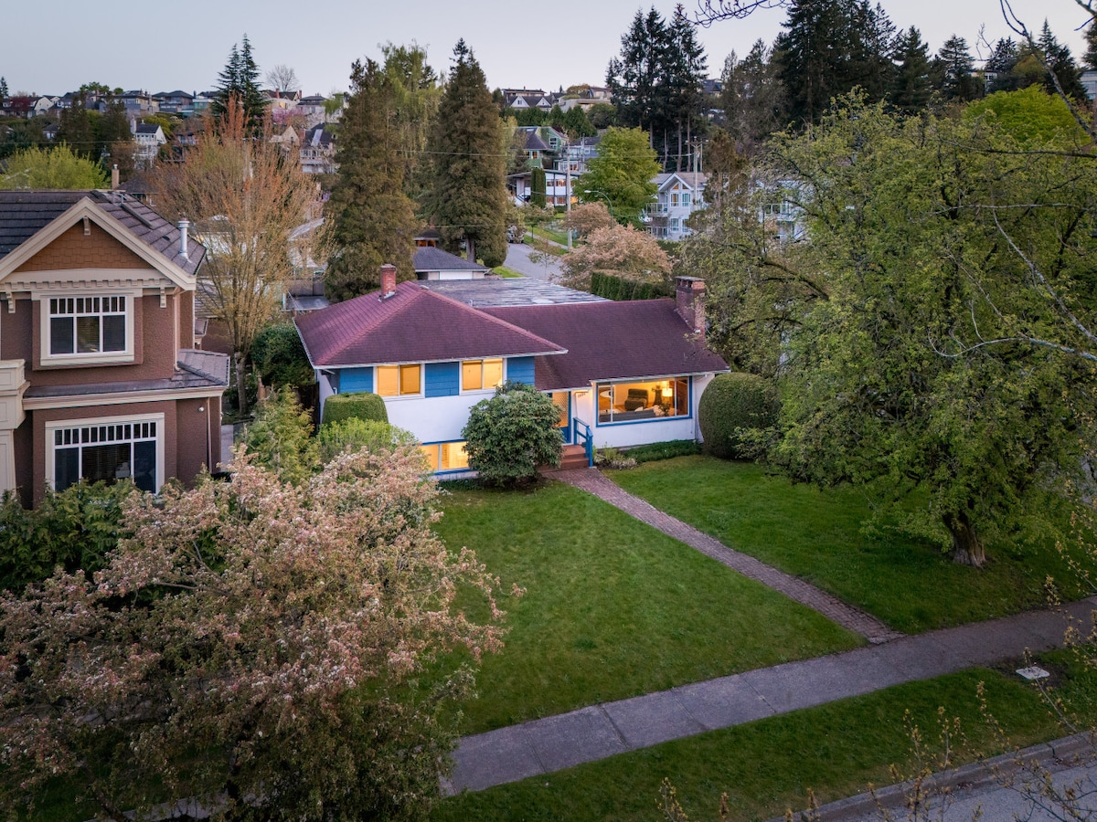 A spacious home is surrounded by well-maintained lawns and trimmed hedges, featuring a welcoming front porch. Nearby trees and blooming flowers contribute to the serene neighborhood. The exterior showcases a combination of light blue siding and a maroon roof, with soft evening light illuminating the front windows.