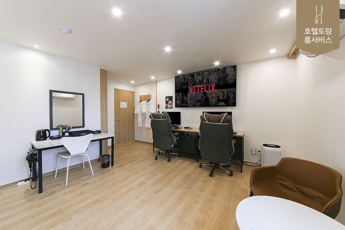 A modern room is presented with light wood flooring and a clean aesthetic. Two workstations equipped with chairs are positioned in front of a large screen displaying Netflix. A small kitchenette with a kettle is visible, alongside a stylish mirror and a closet for convenience.