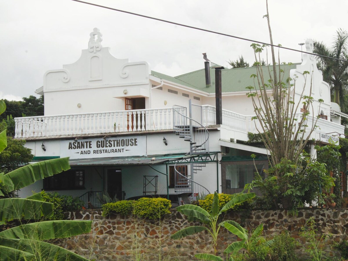 A two-story guesthouse is depicted, accentuated by a white façade and a green roof. A welcoming balcony extends along the front, adorned with decorative elements. Surrounding landscaping features tropical plants and a stone wall, enhancing the inviting appearance of the establishment.