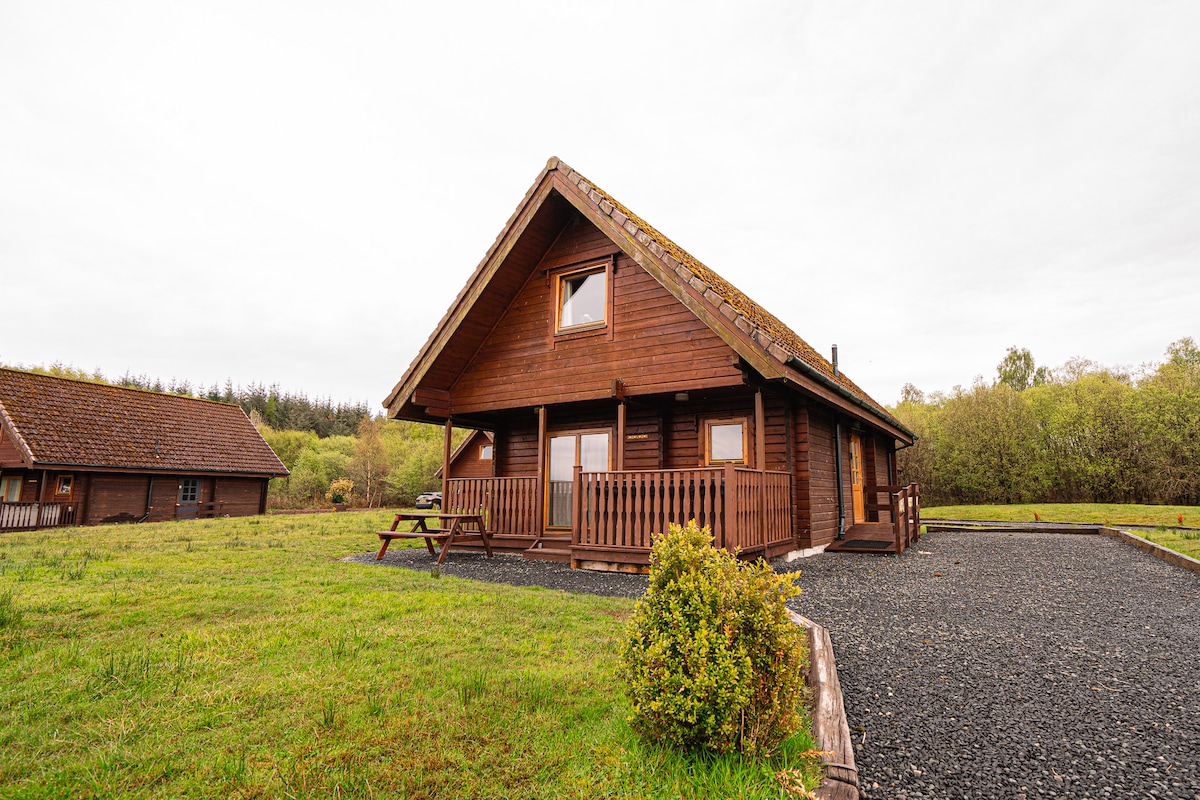 A wooden lodge is set against a backdrop of greenery, featuring a sloped roof and a spacious deck with a railing. The surrounding area includes a manicured lawn and gravel for parking, providing ample space for outdoor activities.