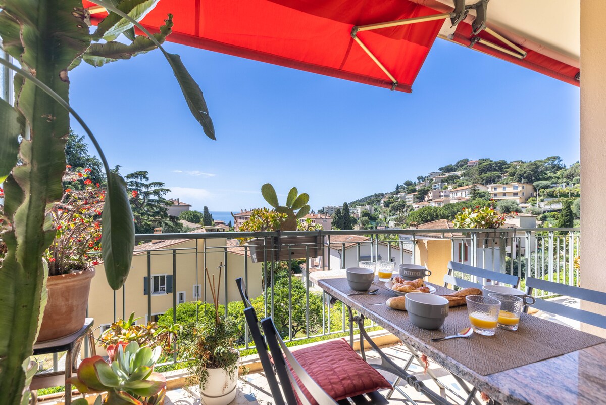 An outdoor balcony is shown, featuring a table set for breakfast with bowls and pastries. Potted plants surround the space, and a red awning provides shade. A clear view of lush greenery and the sea beyond is visible under a bright blue sky.