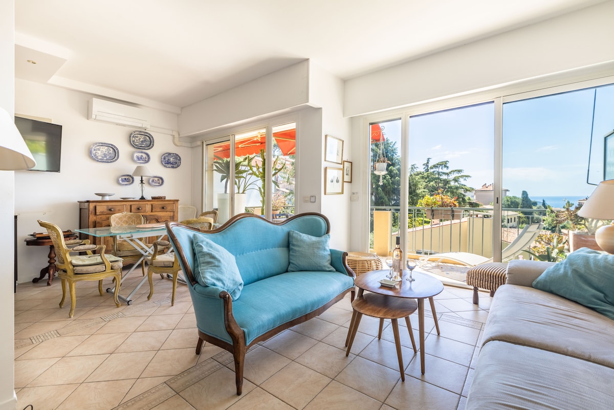 A bright and airy living room features a comfortable seating arrangement with a vintage-style blue sofa and a neutral-toned sofa. Large glass doors provide access to a balcony with a view of the sea, while a dining table and chairs are placed nearby.