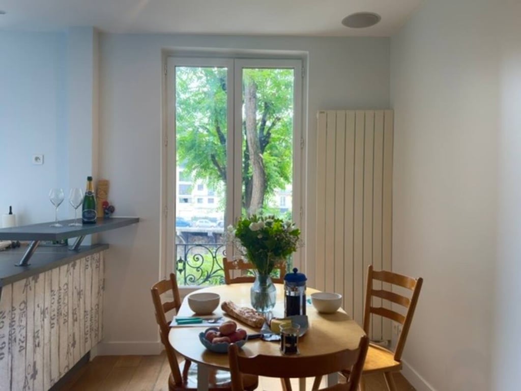 A quaint dining area is depicted, featuring a round wooden table with two bowls, a French press, and fresh produce arranged neatly. Sunlight streams through large windows, illuminating the space, while a green view of trees is visible outside. Simple wooden chairs surround the table.