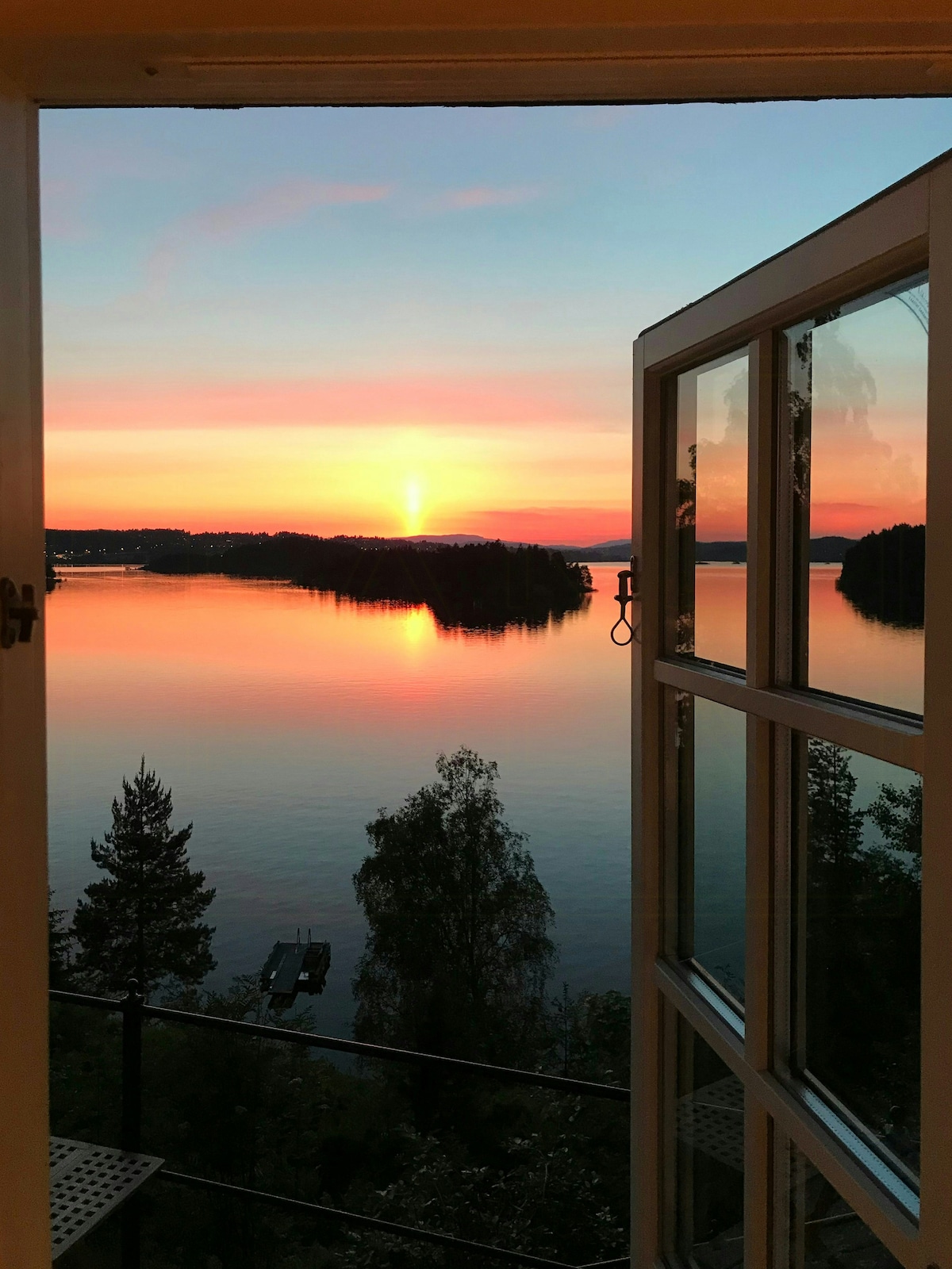 A view of a serene sunset over Steinsfjorden is captured through an open door. The water reflects warm hues of orange and pink, while distant islands are outlined against the twilight sky. The scene invites a sense of calm and connection to nature.