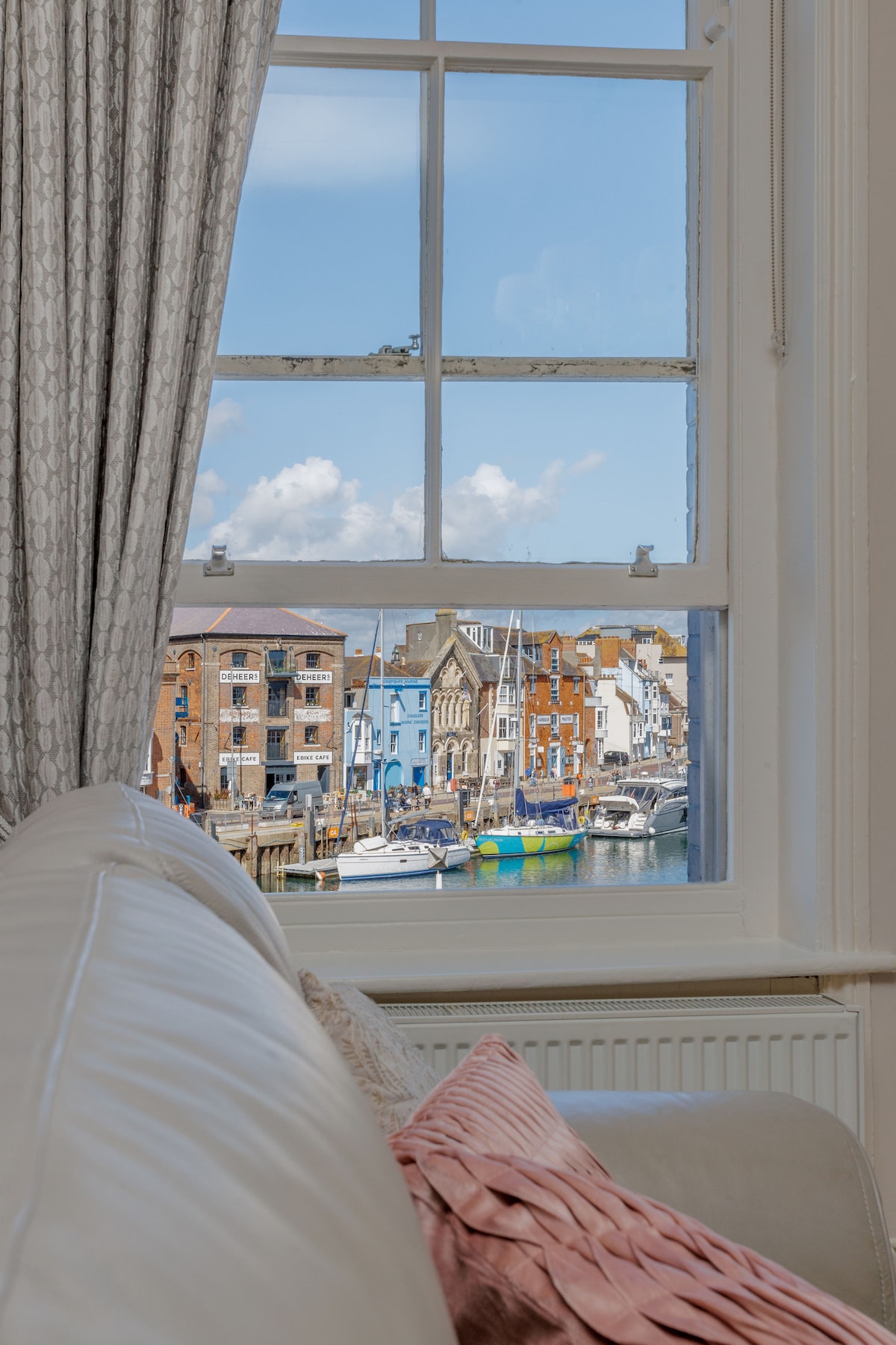 A view of the vibrant harbour is captured through a large window, framed by patterned curtains. Colourful buildings line the waterfront, accompanied by moored boats. A soft white sofa with decorative pillows is positioned in the foreground, providing a cozy viewing spot.