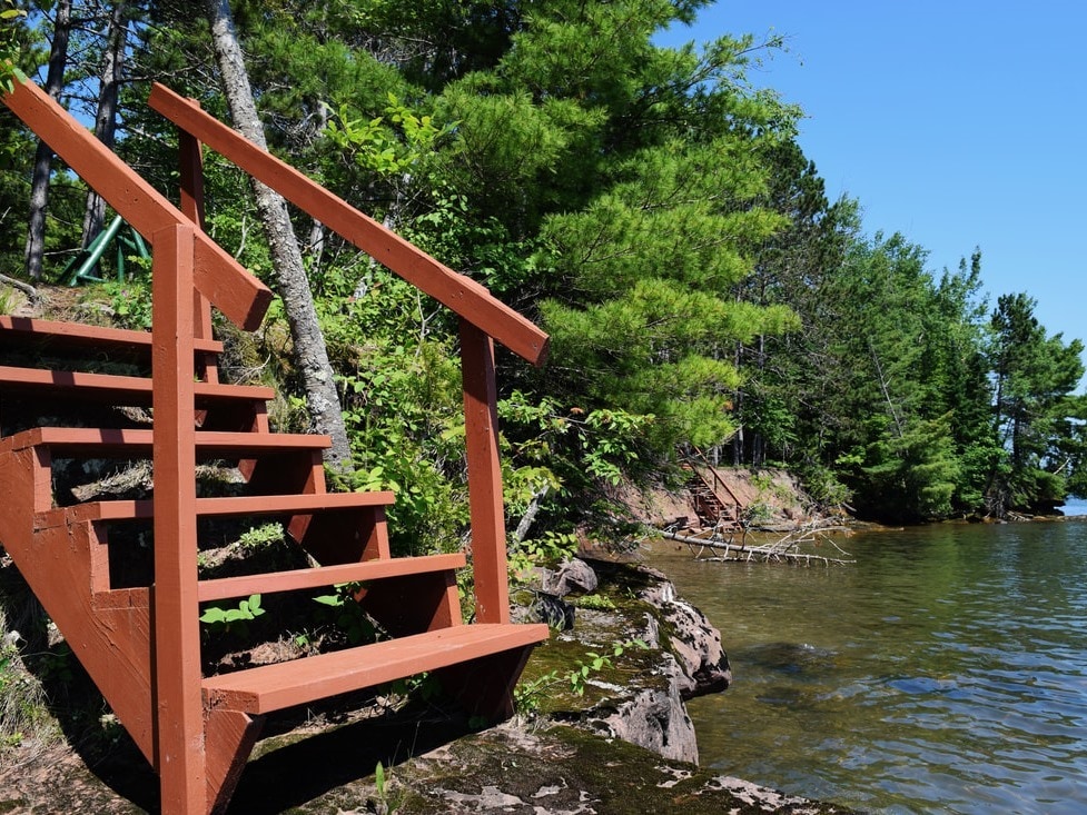 A wooden staircase leads down to the water's edge, surrounded by lush green trees and rocky shoreline. The calm surface of Lake Superior reflects the clear blue sky, providing a serene natural setting. The vibrant wood of the stairs contrasts with the earthy tones of the surrounding landscape.