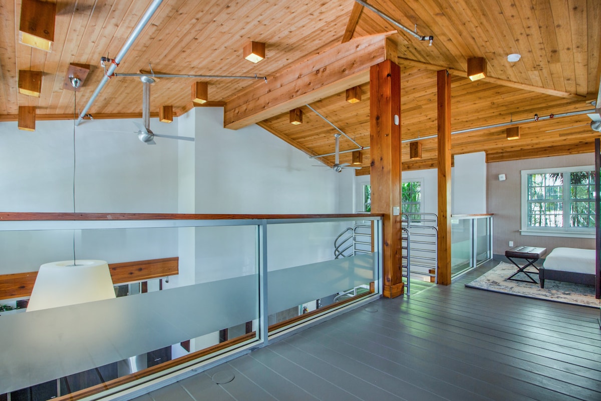 The open loft features a high, wooden ceiling supported by exposed beams. A sleek balcony overlooks the spacious lower level, which is brightly lit by natural light. The flooring combines wood and modern elements, reflecting a blend of comfort and style.