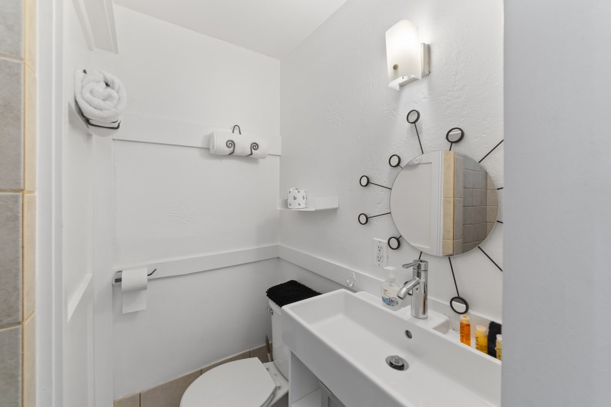 A contemporary bathroom features white walls and a sleek circular mirror mounted above a modern sink. Shelves hold toiletries, while a stylish light fixture illuminates the space. A bath towel and toilet paper are neatly arranged nearby, contributing to the room's inviting and functional design.