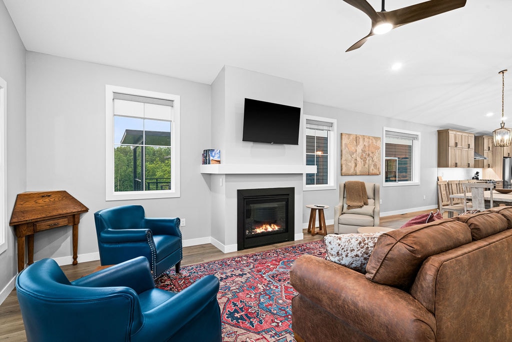 A cozy living area features a brown sofa and blue chairs, arranged around a patterned area rug. A wall-mounted television is positioned above a modern fireplace. Large windows invite natural light, while a dining area and a wooden desk are visible in the background.